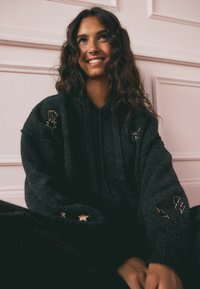 Schwarzer übergroßer Hoodie aus weichem Stoff, mit bestickten Designs: einer Hand und Sternen auf den Ärmeln und dem Wort "Lucky" auf der Brust.