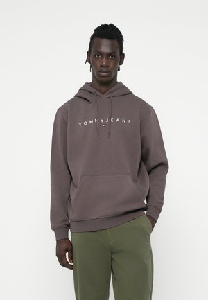 Barna kapucnis pulcsi elülső zsebbel és fehér "TOMMYJEANS" logóval. Zsinóros kapucnival és bordás mandzsettákkal rendelkezik, zöld nadrággal párosítva.