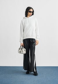 Νέα γυναίκα που φοράει μαύρα γυαλιά ηλίου, λευκή μπλούζα με κουκούλα, μαύρο παντελόνι φόρμας Puma, κρατώντας μια μικρή λευκή τσάντα Puma, στέκεται πάνω σε μπλε χαλί.