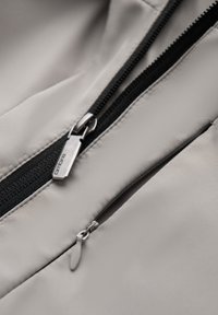 Gros plan de deux fermetures éclair beige sur une veste, l'une partiellement ouverte avec une tirette métallique visible et une étiquette de marque, l'autre plus petite et complètement fermée.