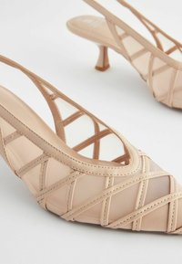 Nude slingback hæle med krydsdesign, gennemsigtigt materiale, spids tå og 2-tommers kitten hæl. Glat, tekstureret finish.