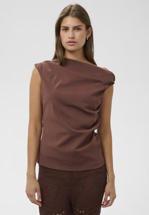 Jeune femme aux longs cheveux bruns et raides, portant un haut marron asymétrique sans manches et un pantalon marron texturé, debout devant un fond uni.