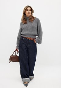 Grau flauschiger Pullover, hoher Kragen, weite dunkelblaue Jeans, geflochtener brauner Gürtel, graue und blaue Manschettendetails, braune Handtasche, spitze graue Schuhe.