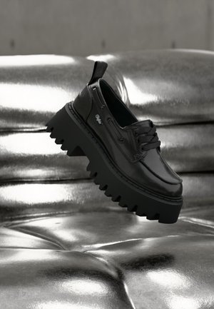 Zwarte leren schoen met dikke zool en dikke veters, rustend op een reflecterend zilverkleurig metalen kussenoppervlak.