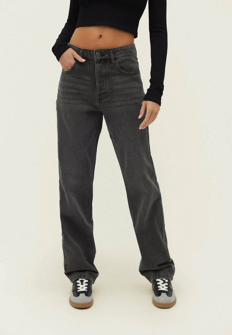 Personne portant un jean noir ample taille haute, un haut noir court à manches longues, et des baskets noir-gris avec des lacets blancs, debout avec une main dans la poche.