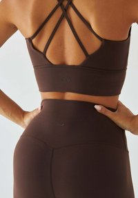 Brązowy zestaw sportowy składający się z krótkiego topu z krzyżowanymi ramiączkami i legginsów z wysokim stanem. Gładka faktura z subtelnym logo na obu elementach.