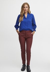 Modrá blůza s dlouhými rukávy a výstřihem do V, s nařasenými rukávy, doplněná burgundskými slim-fit kalhotami; černé kotníkové boty dokončují outfit.