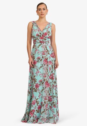 Frau trägt ein ärmelloses, bodenlanges, hellblaues Kleid mit rosa und braunen Blumenmustern und steht vor einem schlichten Hintergrund.