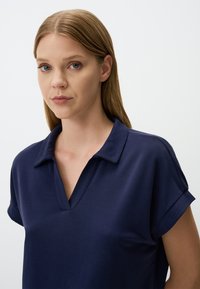 Navyblauwe polo met V-hals en opgerolde korte mouwen, gemaakt van gladde stof, met minimale stiksels en een relaxte pasvorm.