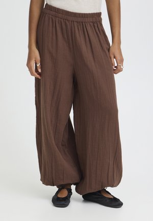 Luźne brązowe spodnie o szerokich nogawkach z elastyczną talią i ściągaczami, noszone z czarnymi płaskimi butami i pierścieniami na obu dłoniach.