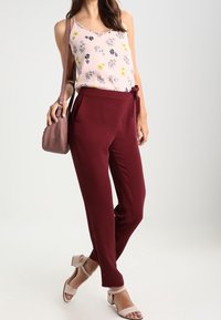 Mouwloze bloemen blouse in lichtroze, gecombineerd met hoge bordeauxrode broeken. Accessoires omvatten een kleine bruine handtas en lichtgekleurde sandalen.