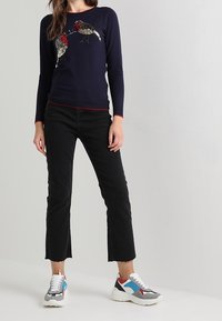 Donna con maglione blu navy con due uccelli di paillettes, pantaloni neri corti e sneakers bianche colorate, in piedi davanti a uno sfondo chiaro e semplice.