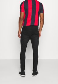 Una persona de espaldas, vestida con una camisa a rayas rojas y azul marino, jeans negros ajustados y zapatillas negras, se encuentra frente a un fondo liso.