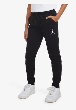 Person iført sorte Jordan joggingbukser med hvidt Jumpman-logo og hvide sneakers, stående med den ene hånd i lommen.