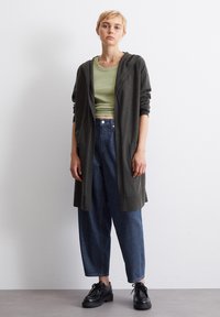 Mørkegrønn lang cardigan med hette, lagd over en lys grønn crop top. Blå løse denimjeans og svarte snøresko fullfører antrekket.
