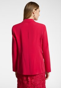 Blazer tailored rosso con una texture liscia, colletto a reverse e spalle strutturate. Presenta una fessura posteriore singola e maniche lunghe.