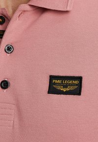Pink polo shirt made of textured fabric, featuring a black label with gold lettering reading "PME LEGEND AMERICAN CLASSIC" and two black buttons.