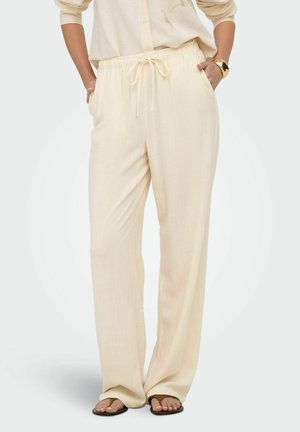 Femme portant un pantalon ample de couleur crème à cordon de serrage avec des poches, assorti à des sandales foncées et un haut à manches longues beige.