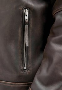 Brown leather jacket featuring a zippered pocket, textured finish, and visible stitching along the seams, adding detail to the design.