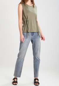 Olivgrön ärmlös topp med insydd midja, kombinerad med ljusblå raka jeans och svarta sandaler med remmar. Neutral bakgrund.