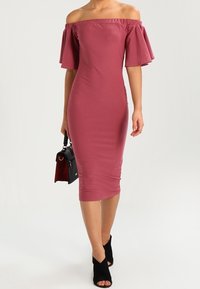 Robe midi ajustée, décolletée épaules dénudées, en tissu mauve avec manches évasées. Associée à des bottines noires et un petit sac à main noir.