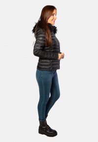 Veste doudoune noire avec un col en fausse fourrure, fermeture éclair à l'avant et design matelassé ; associée à un jean skinny bleu et des bottes noires à lacets.