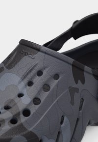 Κοντινή λήψη μαύρου και γκρι μοτίβου καμουφλάζ σε παντόφλα Crocs με τρύπες αερισμού και ρυθμιζόμενο λουράκι στο τακούνι.