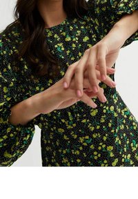 Vestido negro con estampado floral en amarillo y verde, y lunares. Presenta mangas fruncidas y una cintura ceñida. Las manos están ligeramente posadas.