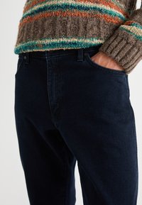 Jeans de color azul oscuro con una forma ajustada, que presentan un bolsillo frontal y una cintura clásica con botón. La textura parece suave y resistente.