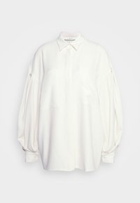 Camicia bianca realizzata in un tessuto liscio, caratterizzata da un colletto classico, due tasche frontali e maniche ampie a sbuffo con polsini a bottoni.