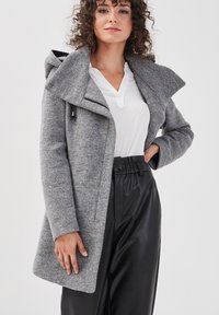 Manteau tricoté gris avec un col oversized et une capuche, doté d'une fermeture éclair à l'avant et d'un tissu texturé. Assorti avec un pantalon noir taille haute.