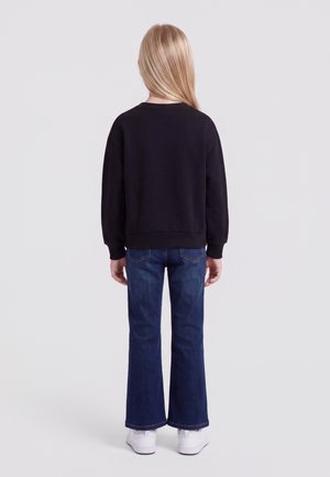 Barn med langt blondt hår iført en sort sweatshirt, blå vidde jeans og hvide sneakers, står med ryggen til på en ensfarvet baggrund.