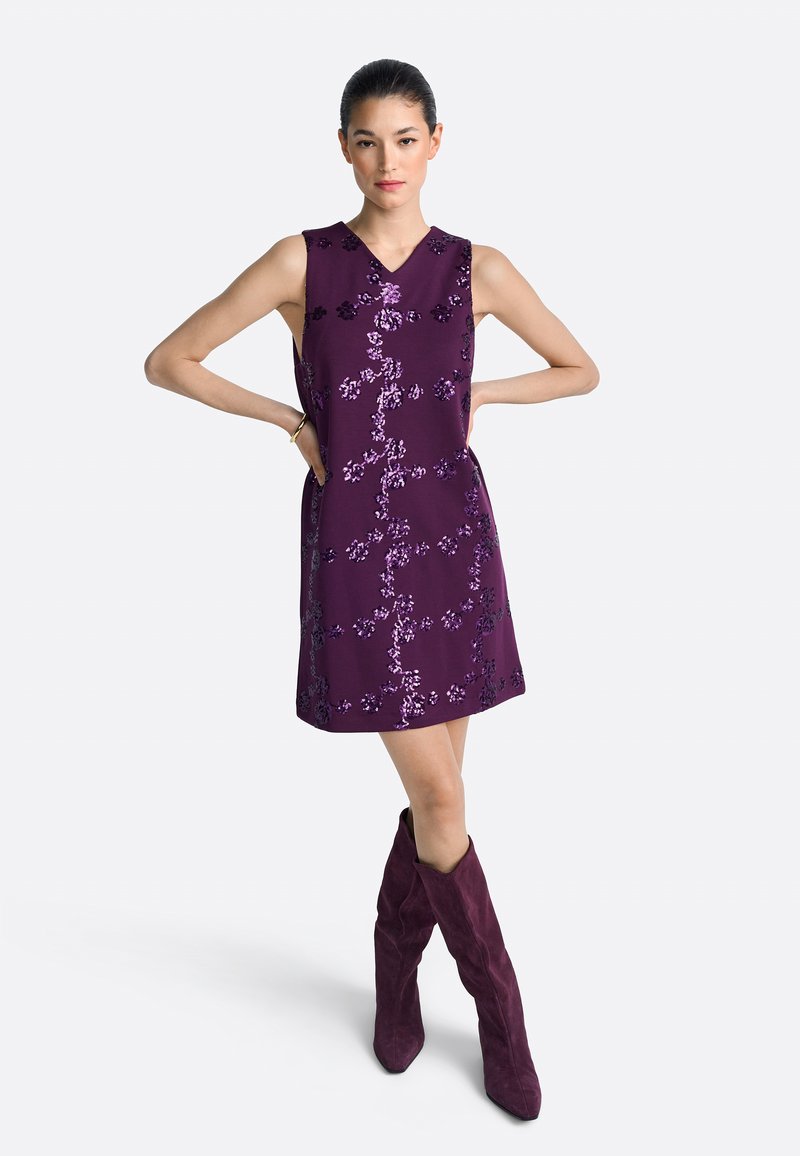 Fioletowa sukienka bez rękawów z kwiatowymi zdobieniami z cekinów, dekoltem w serek i prostą sylwetką; zestawiona z fioletowymi kozakami do kolan.