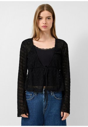 Junge Frau mit glatten hellbraunen Haaren, die ein durchsichtiges schwarzes Langarmshirt über einem schwarzen Tanktop und blauen hoch taillierten Jeans trägt.