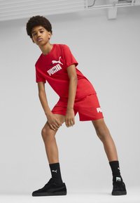 Conjunto de camiseta y pantalones cortos rojos de Puma con un gran estampado de logo, acompañado de zapatos y calcetines negros, sobre un fondo neutro.
