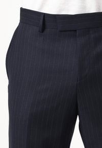 Laivastonsiniset räätälöidyt housut, joissa ohuita pystysuoria pinstripe-raitaita, vyölenkit ja etuosa suljettava, yhdistetty valkoiseen yläosaan.