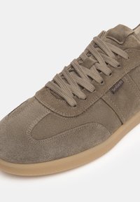 Wildledersneaker in einem gedämpften Olivton, mit einer Gummisohle, strukturiertem Obermaterial und flachen Schnürsenkeln. Die Markenkennung ist an der Seite sichtbar.