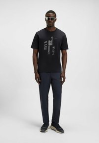 Black short-sleeve t-shirt with a graphic design on the front, paired with dark blue pants and black sneakers. Model wearing sunglasses.