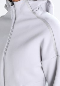 Sudadera con cremallera de color gris claro y cuello alto, con una textura suave y franjas reflectantes a lo largo de los hombros.