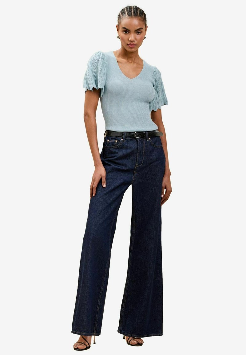 Top tricotat cu dungi, de un albastru deschis, cu slefuri puff, purtat cu blugi din denim cu picioare largi de un albastru închis și o curea neagră. Textură netedă și croială lejeră.