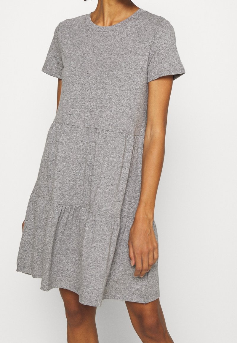 Femme portant une robe grise à manches courtes, longueur genou, à étages, avec un col rond, sur un fond clair uni.