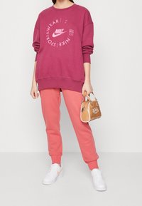 Nike oversize maroon sweatshirt med vit logotyp, tillsammans med rosa joggers och vita sneakers. Håller en tan handväska med logotyp.