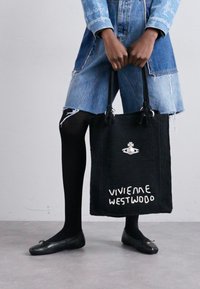 Black woven tote bag with the text "VIVIENNE WESTWOOD" in white. Handles feature black knots. Model wears a denim dress and black tights.