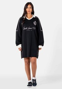 Vrouw in een zwart oversized jurk met lange mouwen, witte kraag, witte manchetten en de tekst "Sixth June Co. Paris", gecombineerd met zwarte Adidas sneakers.
