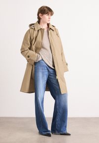 Beige wasserfester Trenchcoat mit Kapuze, beiger Strickpullover und weit geschnittene blaue Jeans. Schwarze spitze Schuhe runden das Outfit ab.