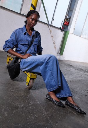 Denim shirt with two pockets, wide-leg blue jeans, black studded flats, and a black shoulder bag, seated on a yellow dolly in an industrial space.