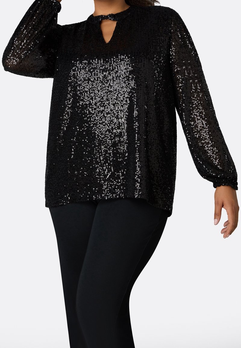 Top nero con paillettes, caratterizzato da maniche lunghe, scollatura a chiave e vestibilità morbida, abbinato a leggings neri. Superficie lucida e strutturata.
