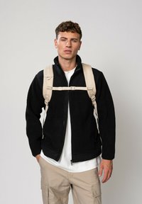 Veste en polaire noire avec un col haut et fermeture zippée, associée à un sac à dos clair porté sur une chemise blanche et un pantalon cargo.