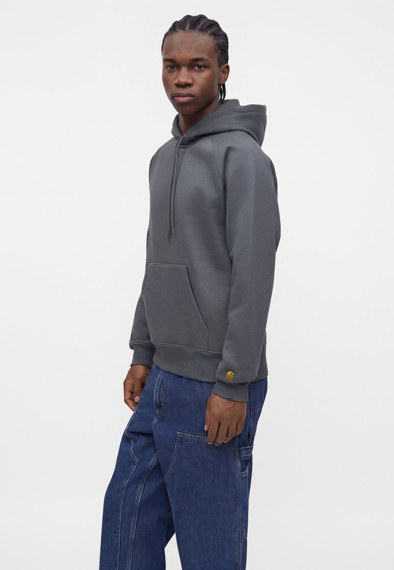 Junger Mann mit geflochtenem Haar, der einen grauen Kapuzenpullover und weite blaue Jeans trägt, steht vor einem schlichten weißen Hintergrund.