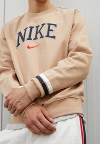 Beige sweatshirt med "NIKE"-logotyp i marinblå och röd brodyr, med randiga muddar i marinblått, vitt och beige. Mjuk tygtextur.